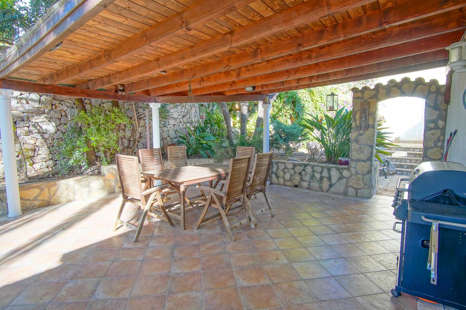 Espace repas extérieur abrité par une pergola en bois, comprenant un barbecue, des murs en pierre naturelle et du mobilier de jardin rustique.