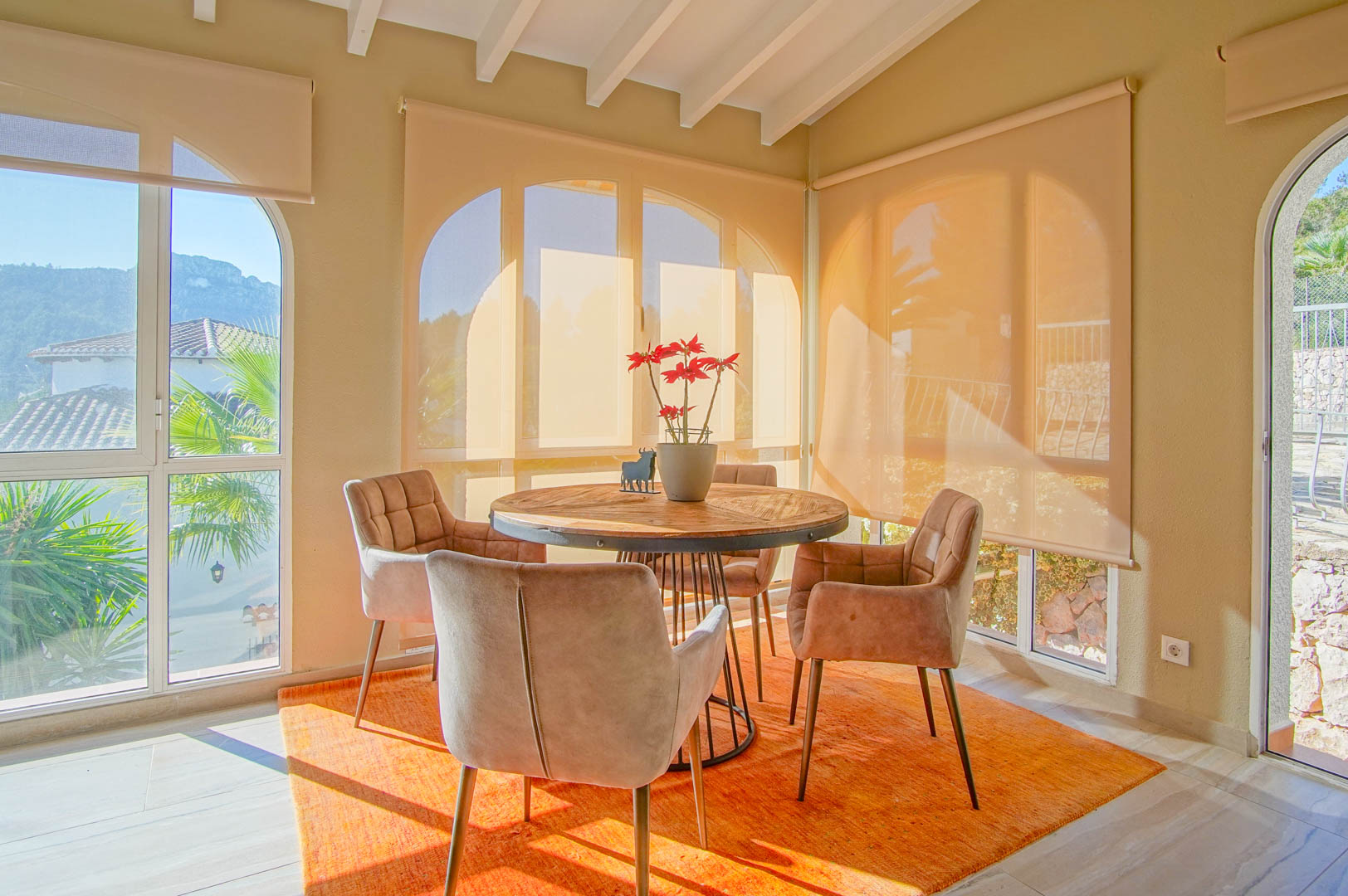 Espace repas lumineux avec table ronde en bois, poutres blanches apparentes et grandes fenêtres offrant une vue sur les montagnes.