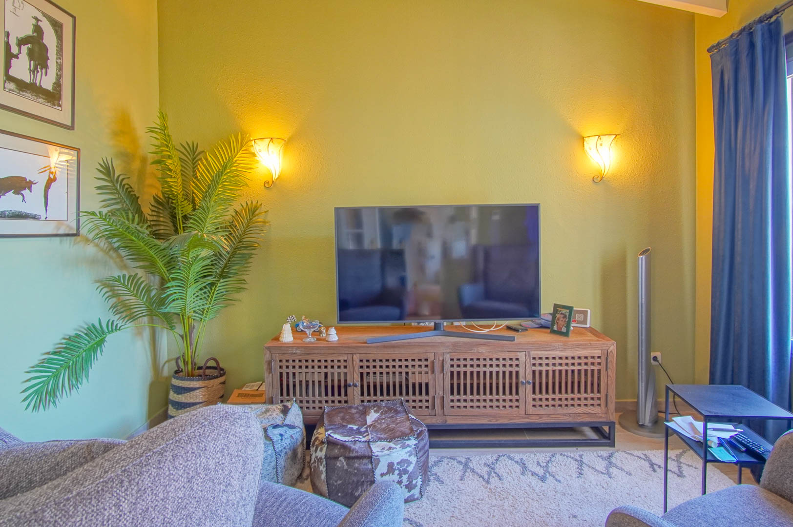 Espace salon avec meuble TV en bois sculpté, éclairage d'ambiance par appliques murales, plante verte et poufs en peau de bête.