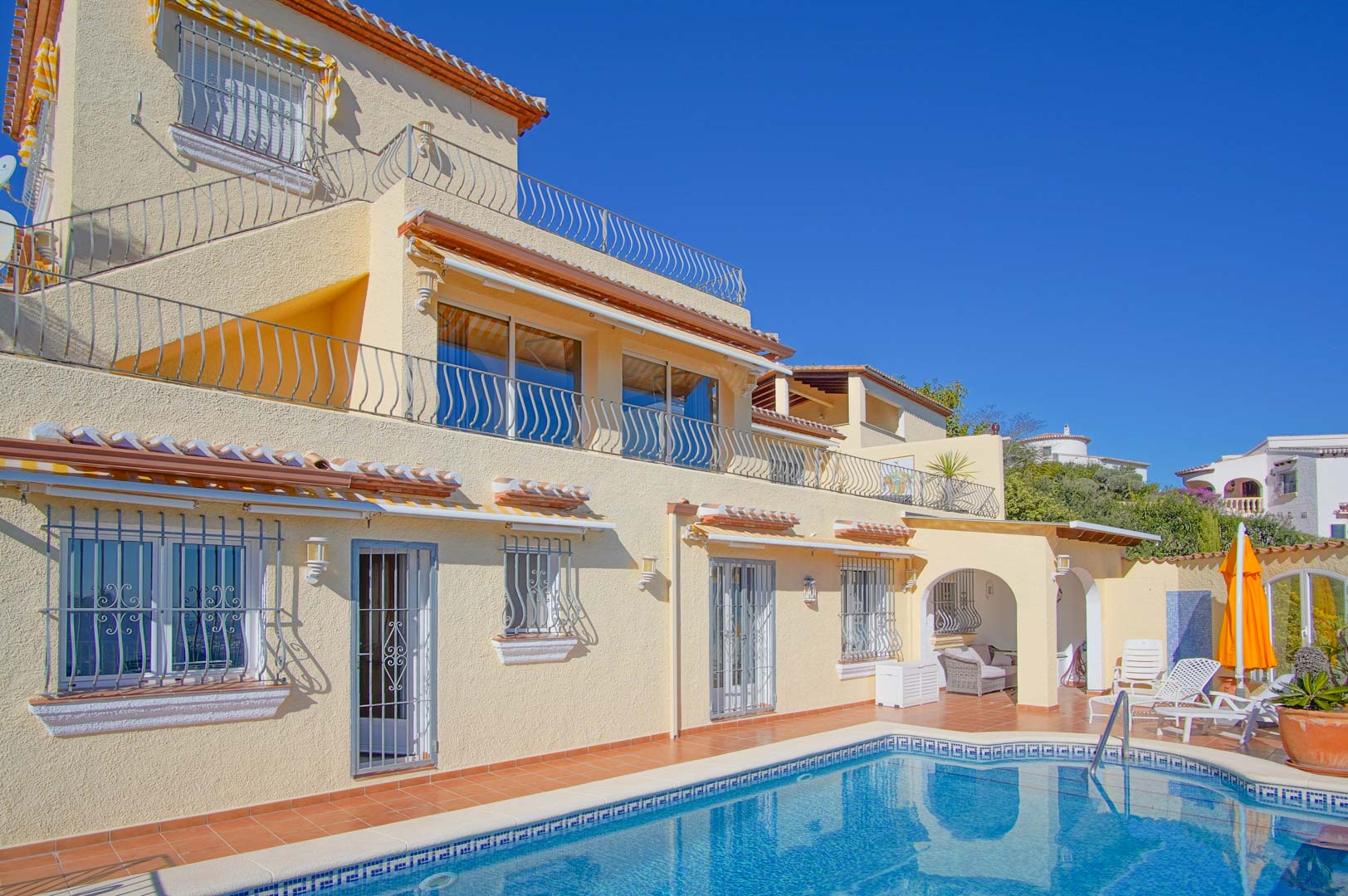 Façade de trois niveaux avec piscine en mosaïque bleue, vastes terrasses avec garde-corps en fer forgé et solarium en terre cuite.