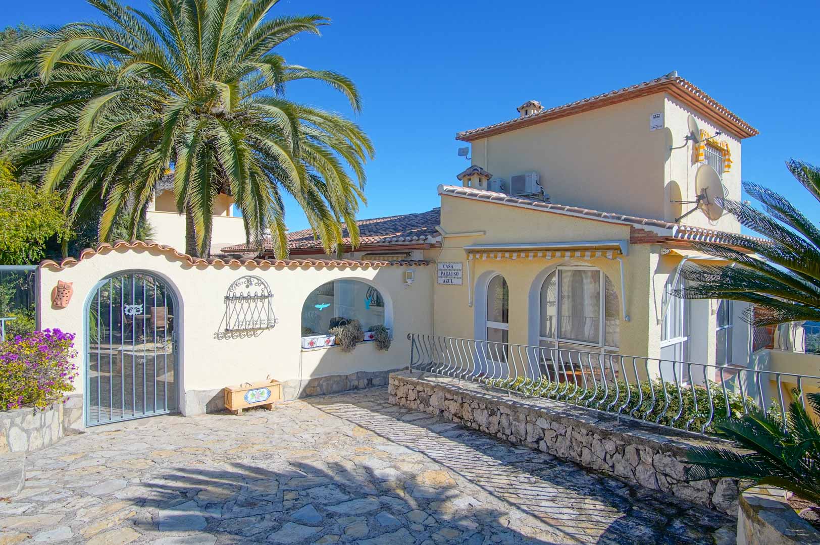 Façade principale de la villa avec entrée pavée, murs en pierre naturelle, arches décoratives et végétation méditerranéenne sous un ciel dégagé.
