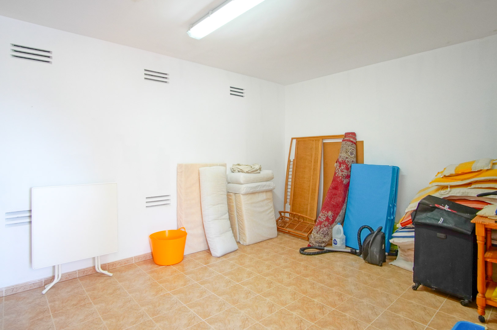 Pièce de rangement fonctionnelle avec murs blancs, grilles de ventilation et sol en carrelage. Un espace polyvalent éclairé par un plafonnier fluorescent.