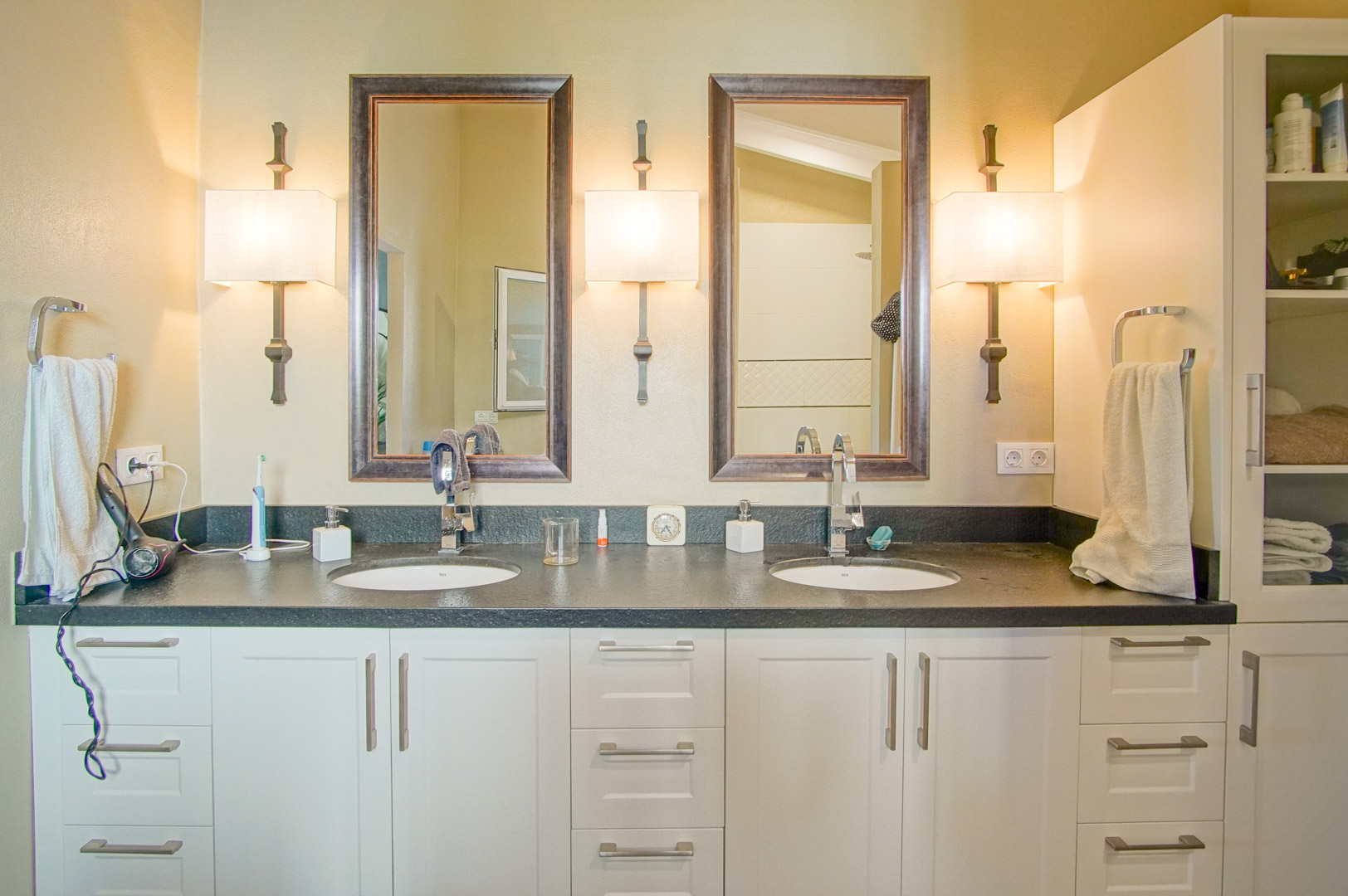 Salle de bain avec double vasque, plan de travail en pierre sombre et meubles blancs. Deux miroirs encadrés sont éclairés par trois appliques murales classiques.