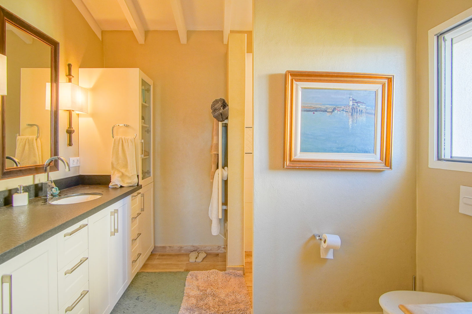 Salle de bain avec plan de travail sombre, poutres en bois apparentes et meubles blancs. L'espace dispose d'un éclairage chaleureux et d'une fenêtre latérale.