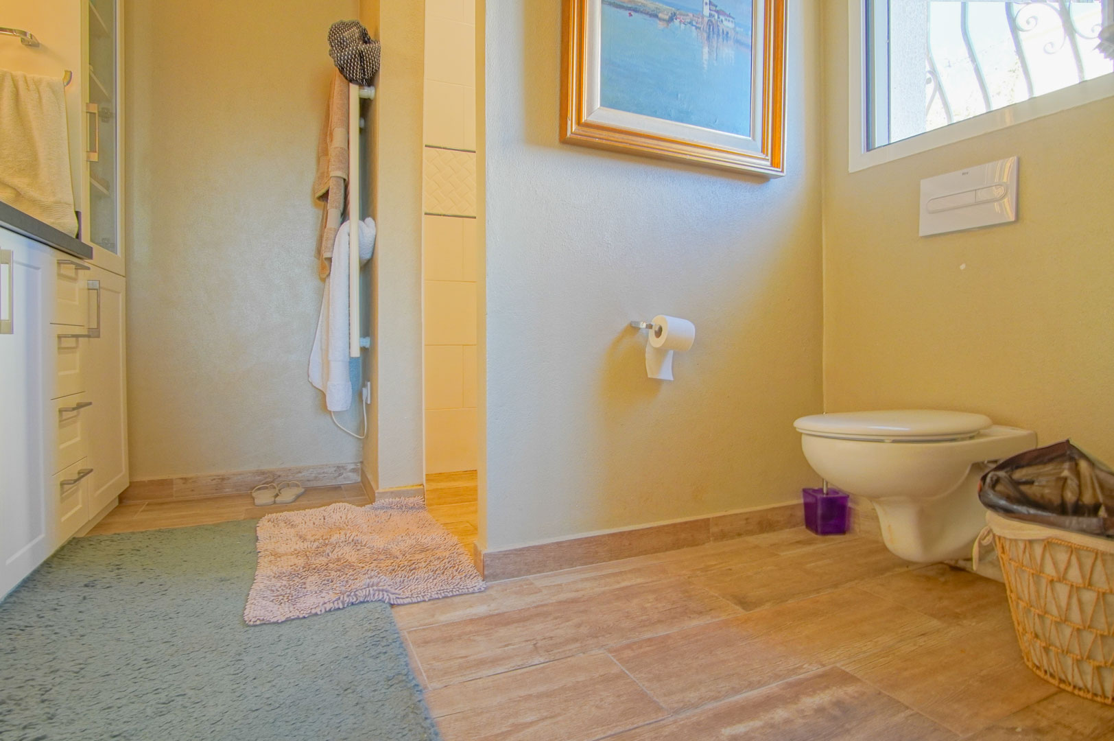 Salle de bain avec WC suspendu, sol en grès cérame imitation bois et meubles blancs. Comprend un radiateur sèche-serviettes et un accès à la douche à l'italienne.