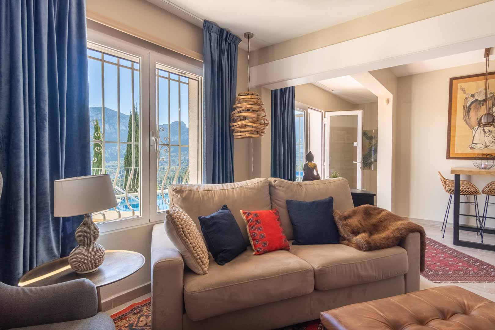 Salon lumineux avec canapé beige, rideaux en velours bleu et fenêtres offrant une vue sur la montagne. L'espace s'ouvre sur un coin bar avec tabourets en osier.