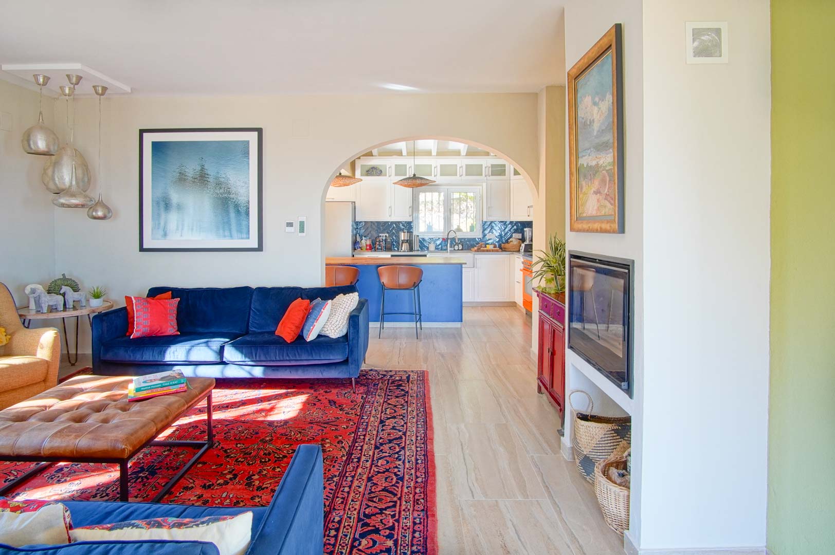 Salon lumineux avec canapés en velours bleu et cheminée intégrée, s'ouvrant par une arche sur une cuisine avec îlot central bleu et plan de travail en bois.