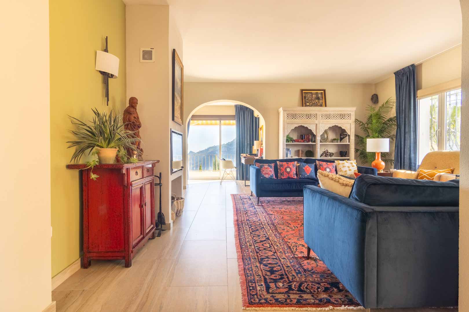 Salon lumineux avec canapés en velours bleu, tapis oriental et arche ouvrant sur une vue montagneuse. Comprend un buffet blanc sculpté et un sol en grès cérame.