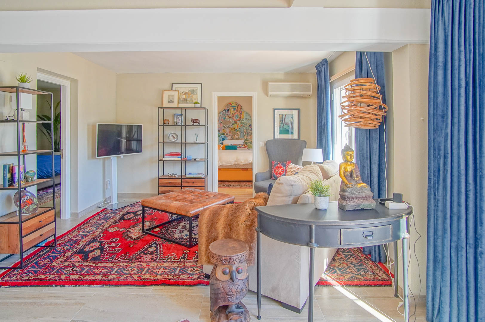 Salon lumineux avec tapis oriental rouge, pouf en cuir capitonné et mobilier éclectique, offrant une vue sur une chambre adjacente décorée.