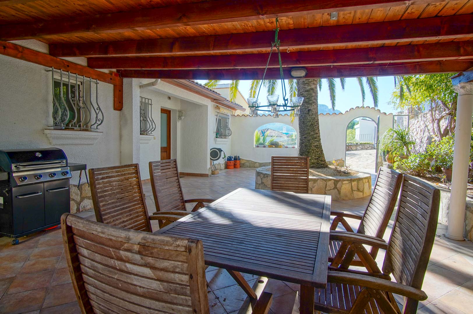 Terrasse couverte avec plafond en poutres de bois, coin repas extérieur, barbecue à gaz et patio avec palmier et murets en pierre.
