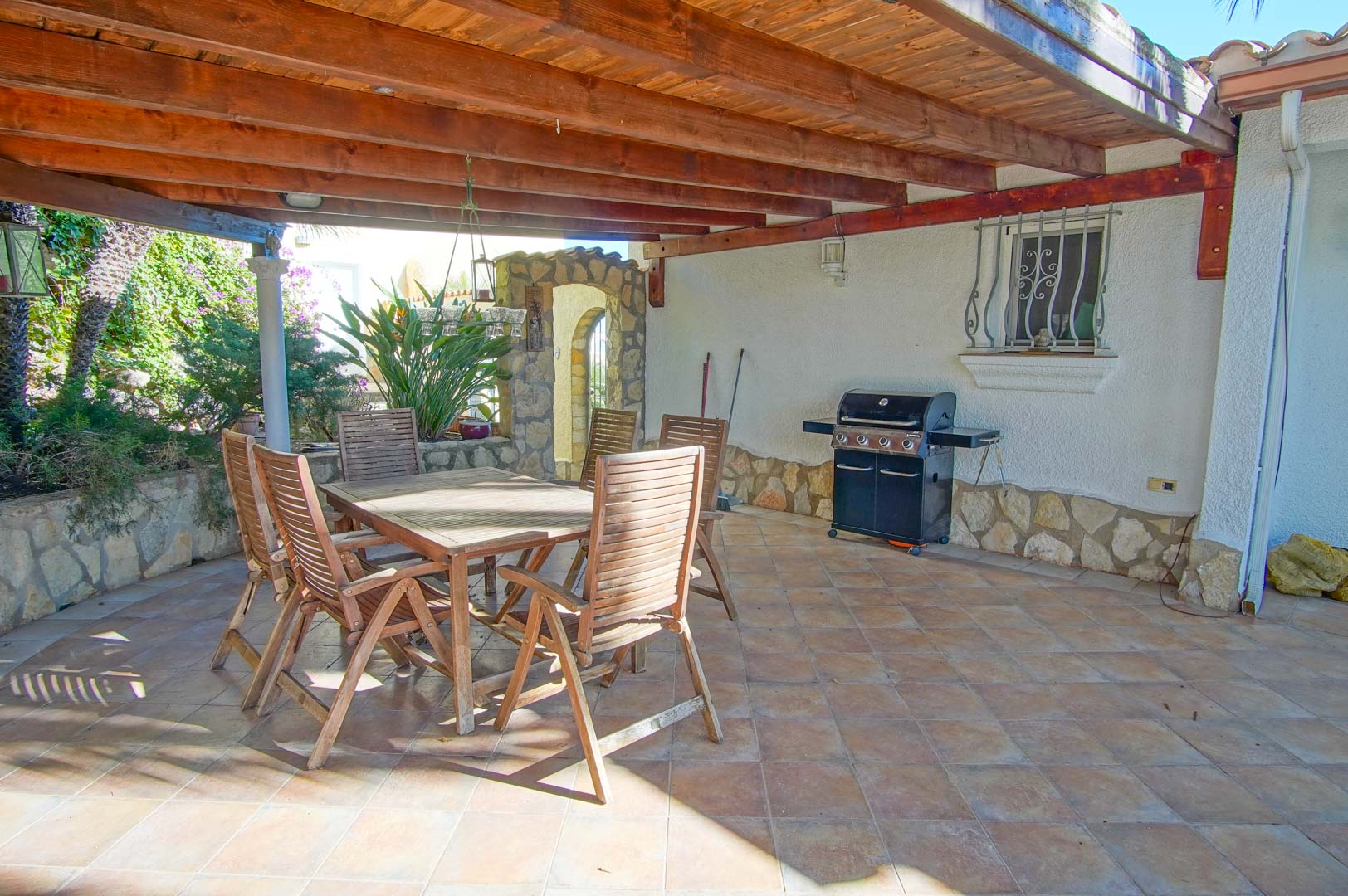 Terrasse couverte avec poutres en bois, espace repas extérieur et barbecue à gaz, complétée par un sol en carrelage et des finitions en pierre.