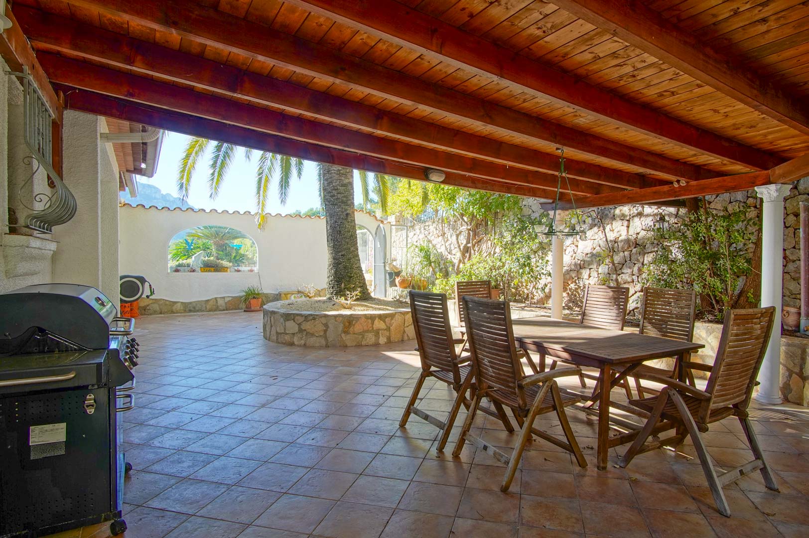 Terrasse couverte avec poutres en bois, salle à manger extérieure, barbecue et jardinière circulaire en pierre abritant un palmier, entourée de murs en pierre.