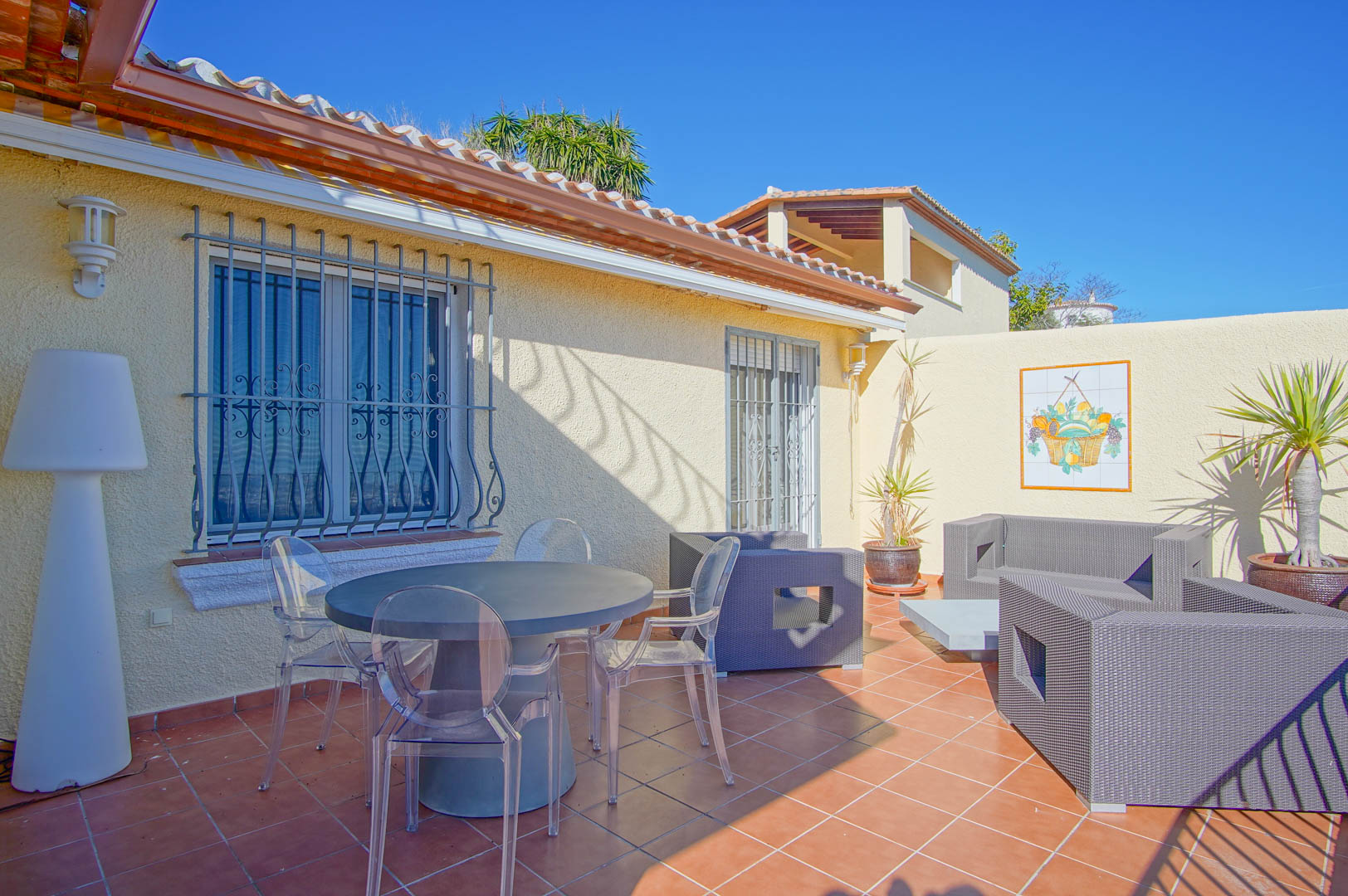 Terrasse extérieure avec carrelage en terre cuite, table ronde avec chaises transparentes et un salon de jardin en résine tressée grise.
