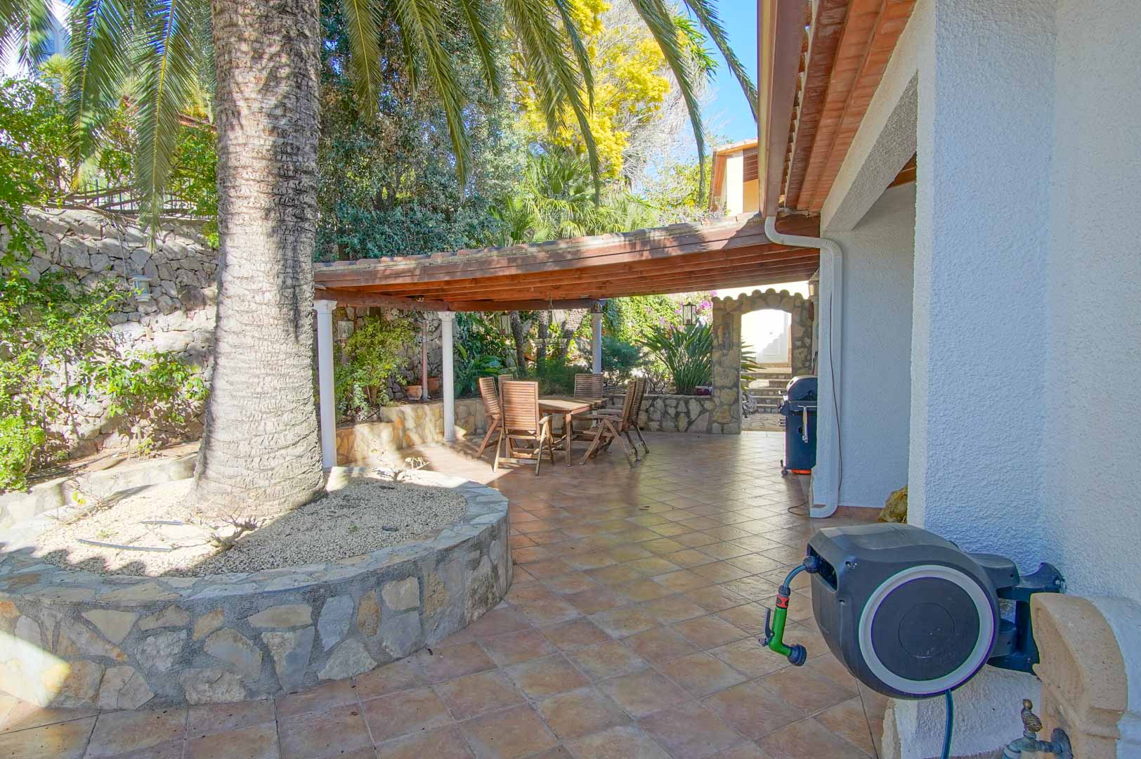 Terrasse extérieure avec espace repas sous une pergola en bois, murs en pierre naturelle et une grande palme entourée d'un sol en carrelage.