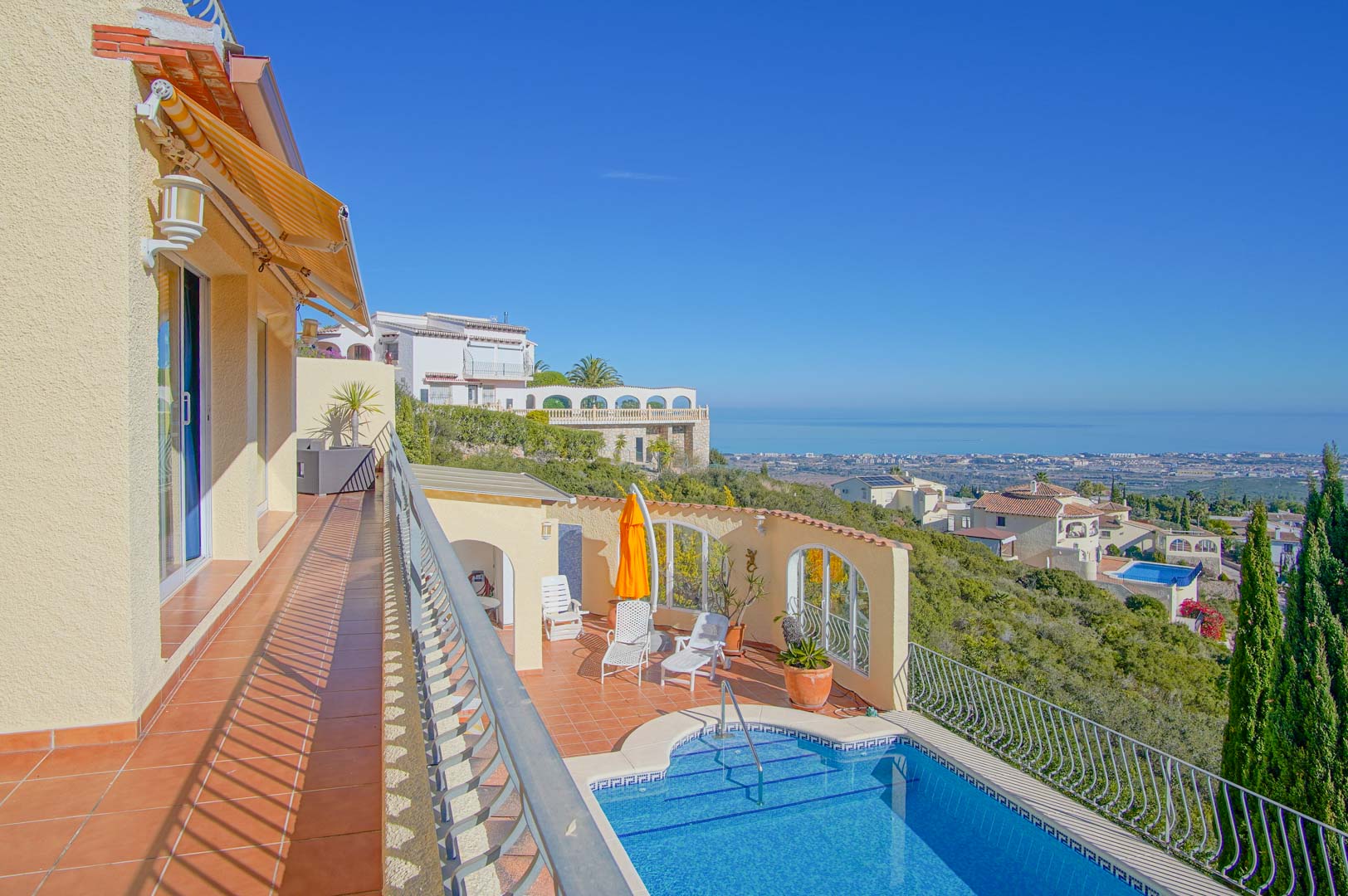 Terrasse supérieure avec carrelage en terre cuite et store, surplombant l'espace piscine et offrant une vue panoramique sur le littoral méditerranéen.