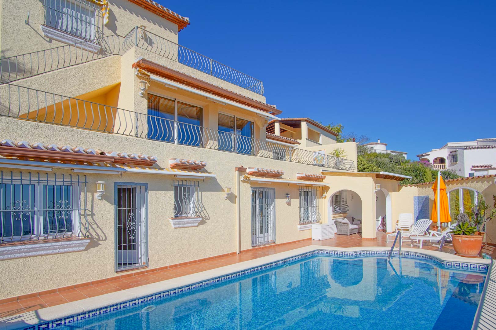 Vue extérieure de la villa avec piscine privée, terrasses en terre cuite et façade sur trois niveaux. Comprend un espace détente sous arche et des garde-corps en fer forgé.