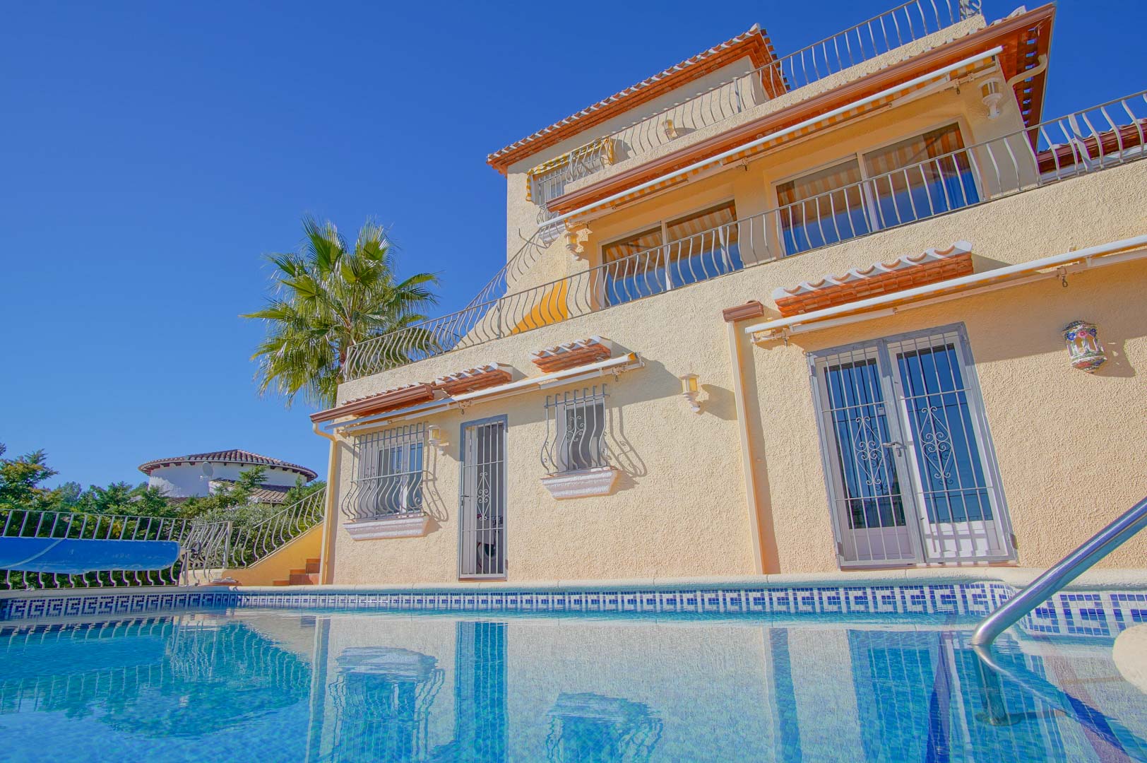 Vue extérieure de la villa sur trois niveaux depuis la piscine, mettant en valeur les terrasses superposées et les détails en fer forgé de la façade.