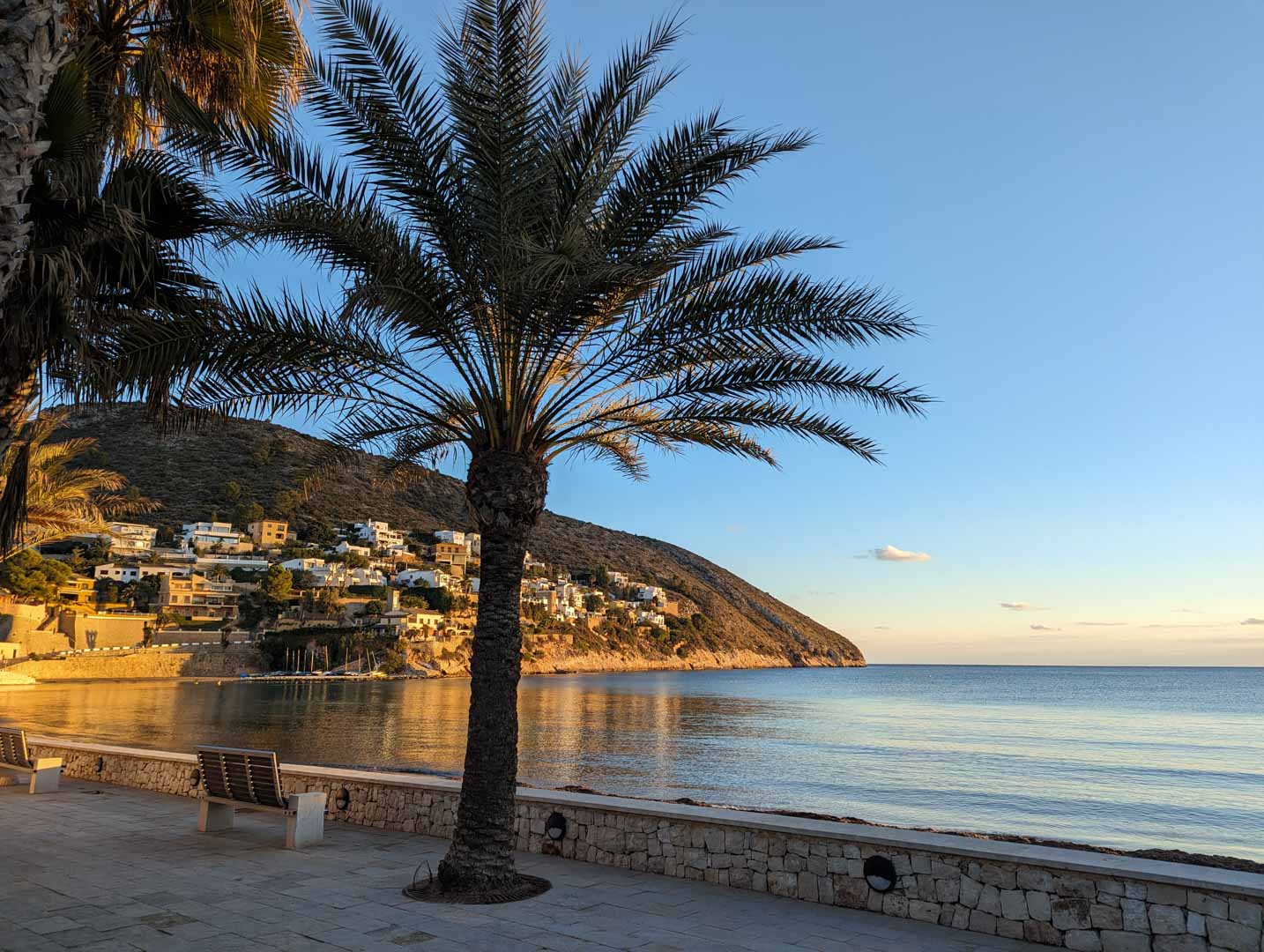 Promenade côtière pavée à Moraira avec palmiers et bancs, offrant une vue sur la mer Méditerranée et les villas nichées sur la colline au coucher du soleil.