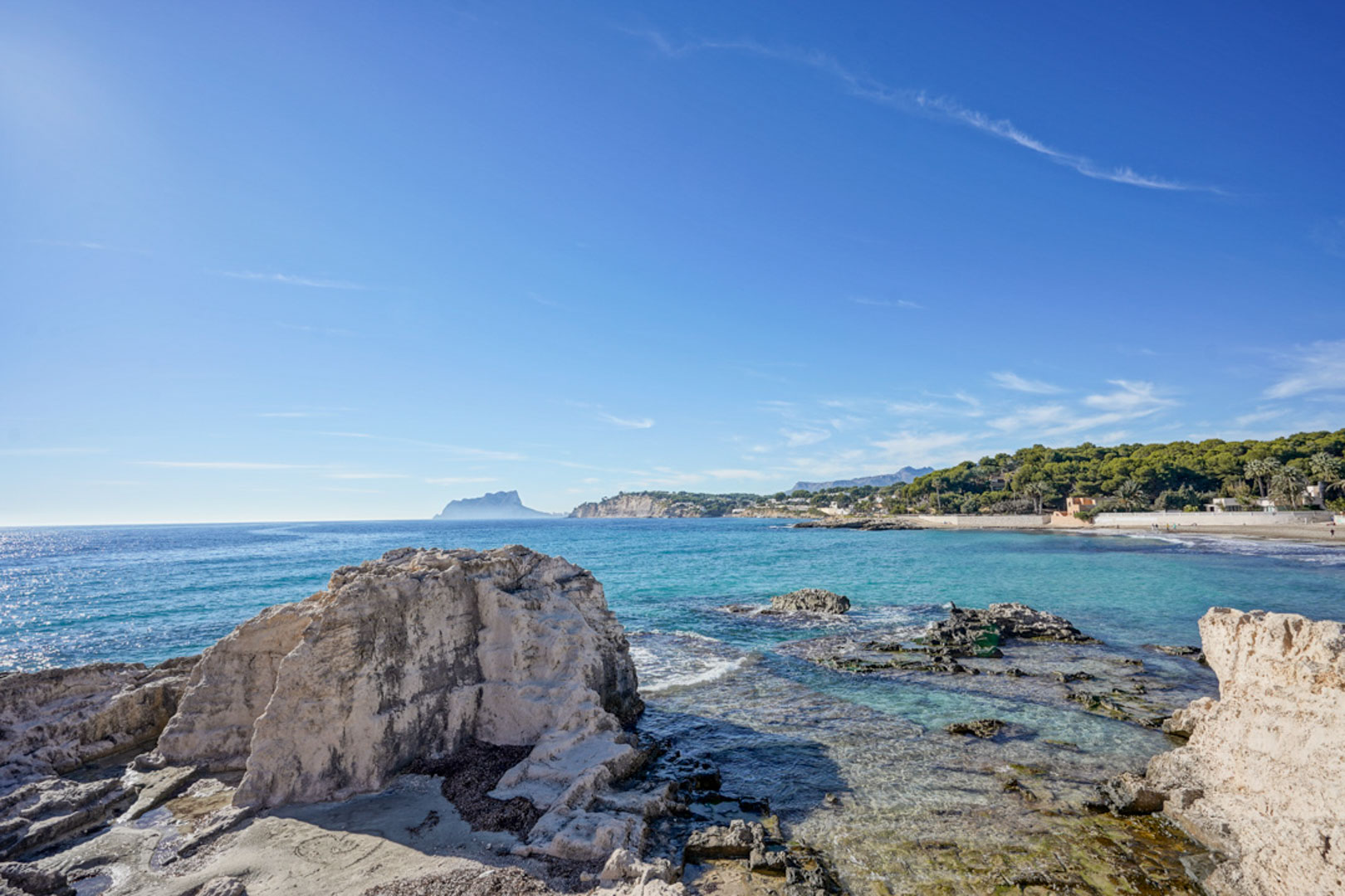 Vue panoramique de la côte de Moraira avec ses formations rocheuses naturelles, ses eaux turquoise cristallines et la silhouette du Peñón d'Ifach à l'horizon.