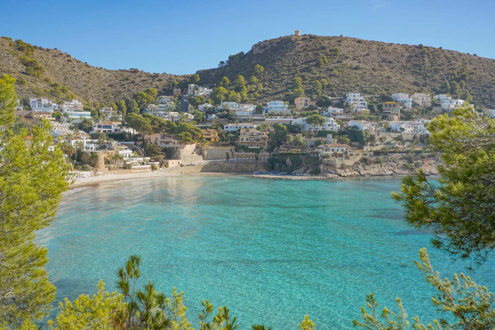 Vue panoramique sur la baie d'El Portet à Moraira, avec ses eaux turquoise cristallines, sa plage de sable et ses collines parsemées de villas méditerranéennes.