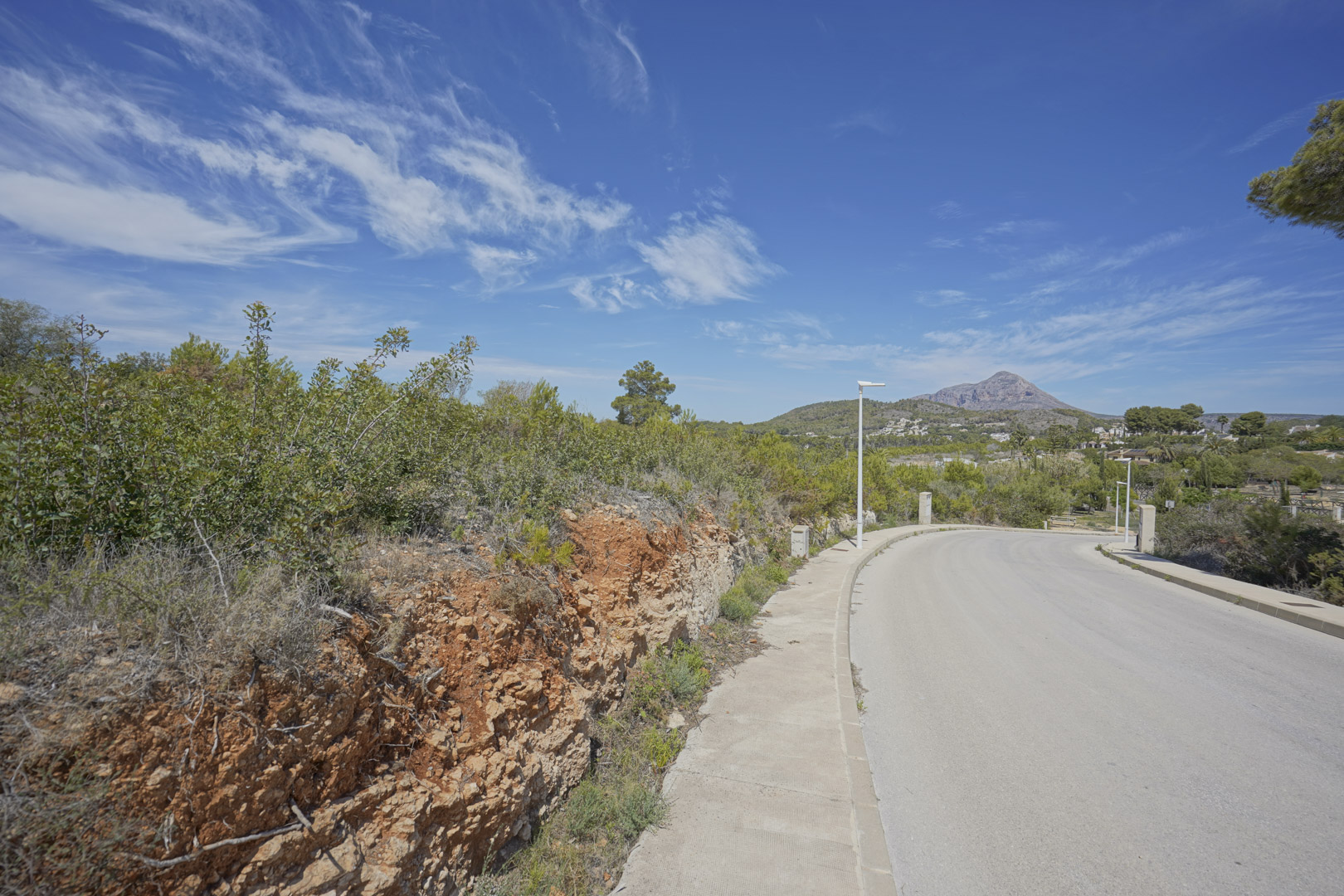 Vue de la route d'accès dans le quartier résidentiel de Valsol à Jávea, avec le mont Montgó en arrière-plan et une végétation méditerranéenne typique.