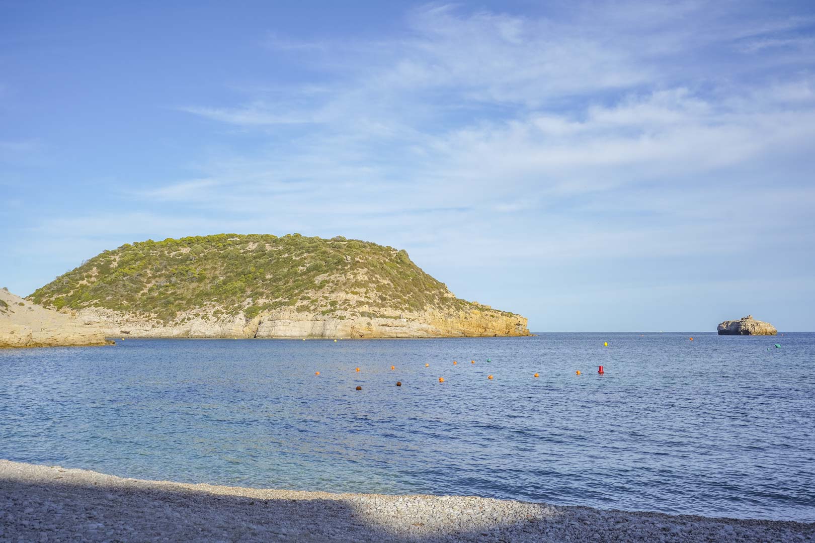 Vue panoramique d'une plage de galets à Jávea, avec les eaux cristallines de la Méditerranée et des falaises côtières sous un ciel dégagé.