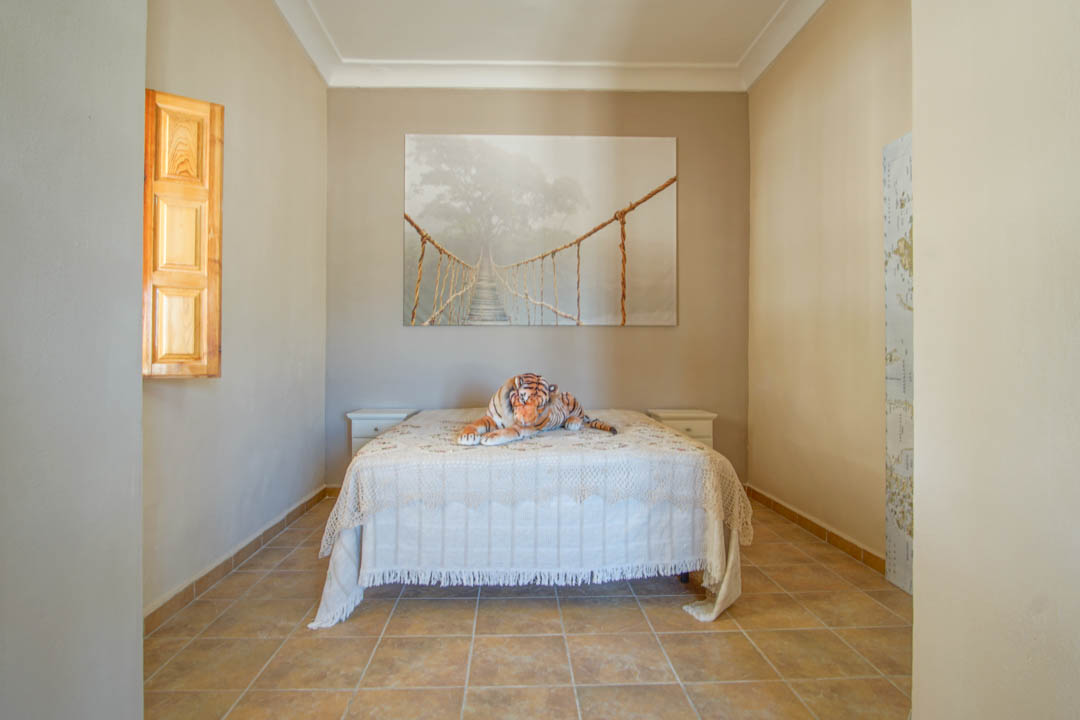 Chambre avec sol en grès cérame, lit double encadré de tables de chevet blanches et fenêtre avec volets intérieurs en bois naturel.