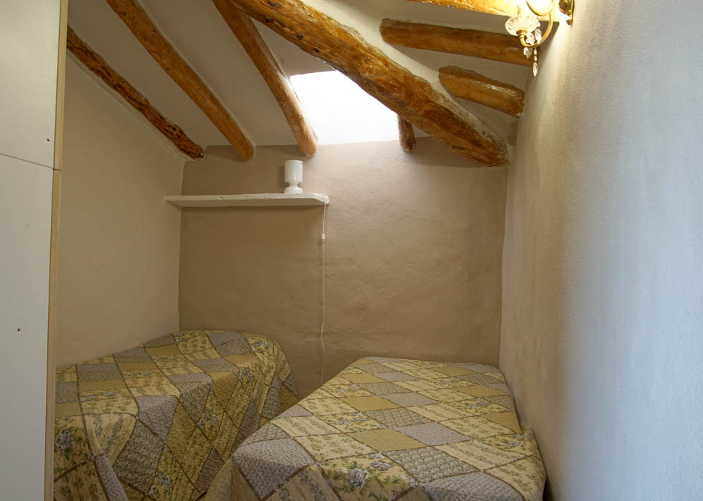 Chambre rustique avec plafonds mansardés, poutres en bois apparentes et lucarne. Elle dispose de deux lits simples et de murs aux finitions en plâtre naturel.