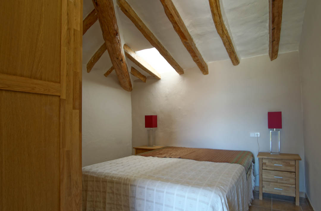 Chambre rustique avec poutres en bois apparentes, lucarne apportant une lumière naturelle et murs en plâtre blanc. Équipée de tables de chevet en bois et lampes rouges.