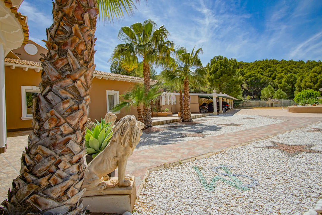 Entrée extérieure avec statues de lions en pierre, palmiers et allée en gravier décorée de mosaïques menant à un abri de voiture couvert bordé par une forêt de pins.