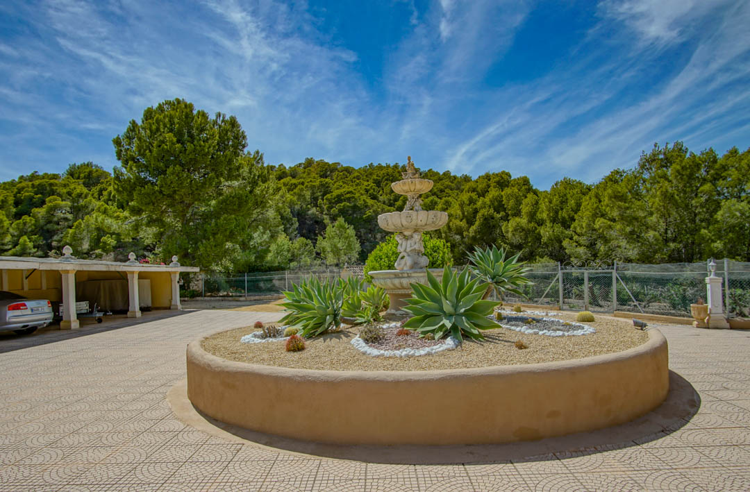 Entrée principale avec fontaine classique à trois niveaux et jardinière circulaire d'agaves, à côté d'un abri pour voitures et d'une pinède.