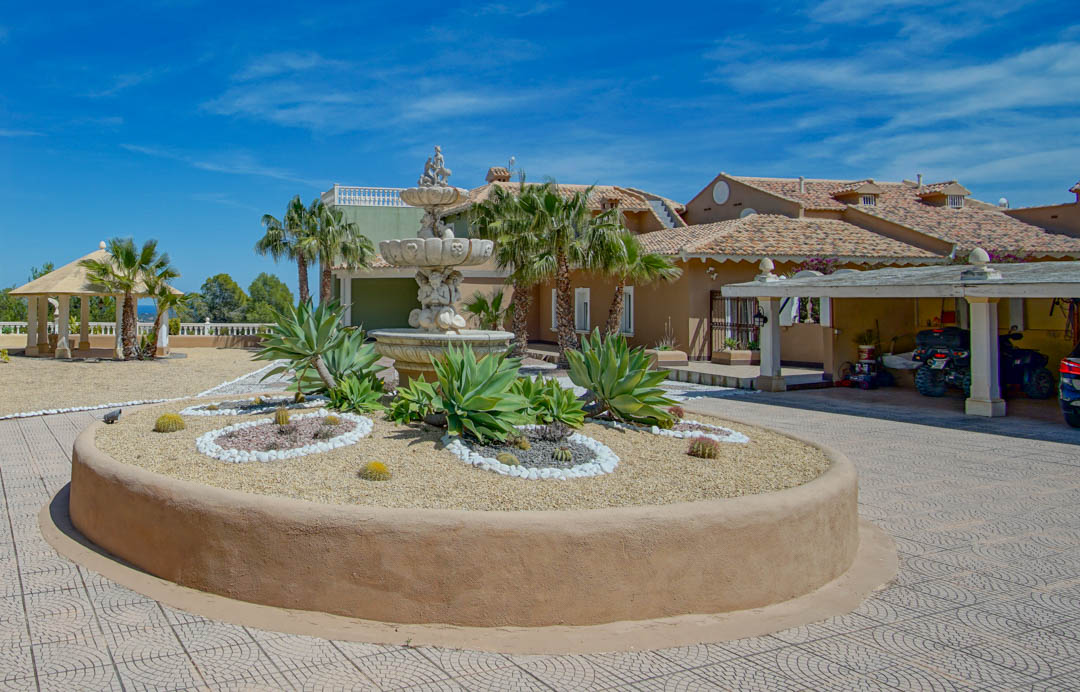 Entrée principale avec fontaine ornementale en pierre, parterre circulaire d'agaves et de cactus, abri de voiture couvert et pavillon de jardin.