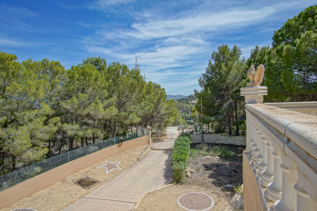 Entrée sécurisée avec allée pavée, balustrade en pierre ornée de sculptures et vue sur la pinède environnante sous un ciel dégagé.