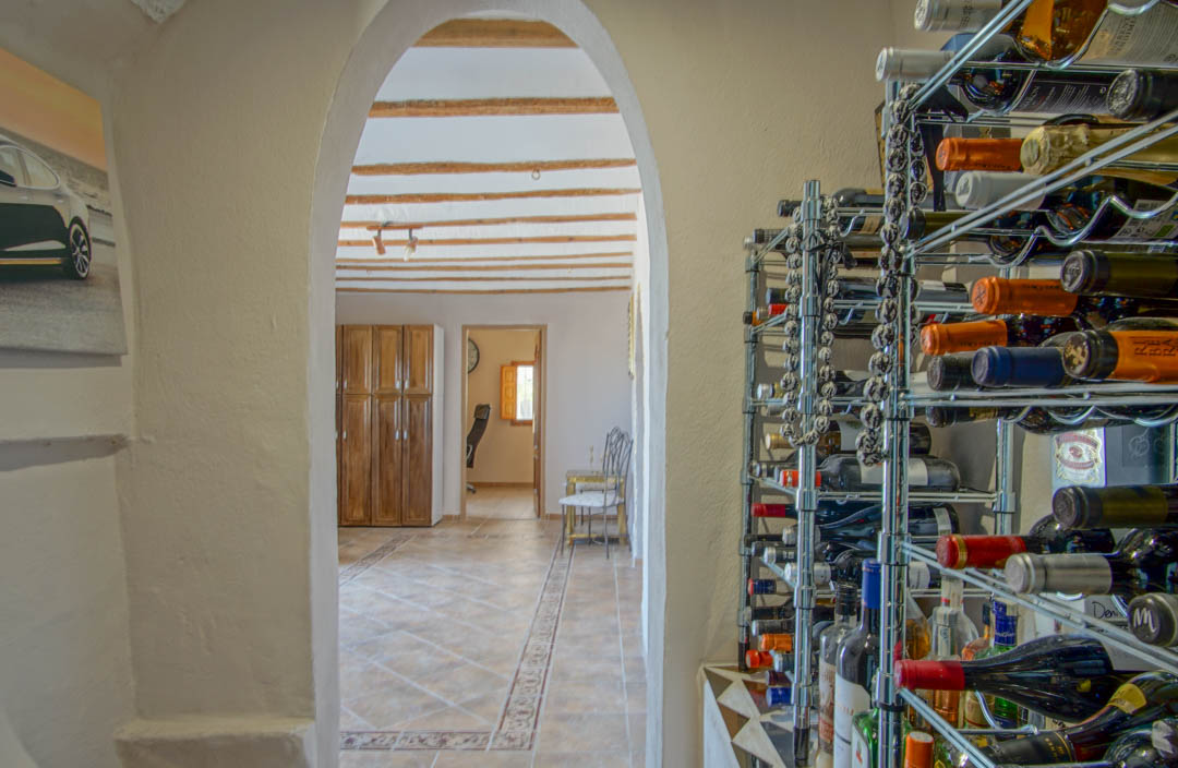 Espace de stockage de vin avec étagères chromées, offrant une vue via une arche sur une pièce aux poutres apparentes et sols en carrelage traditionnel.