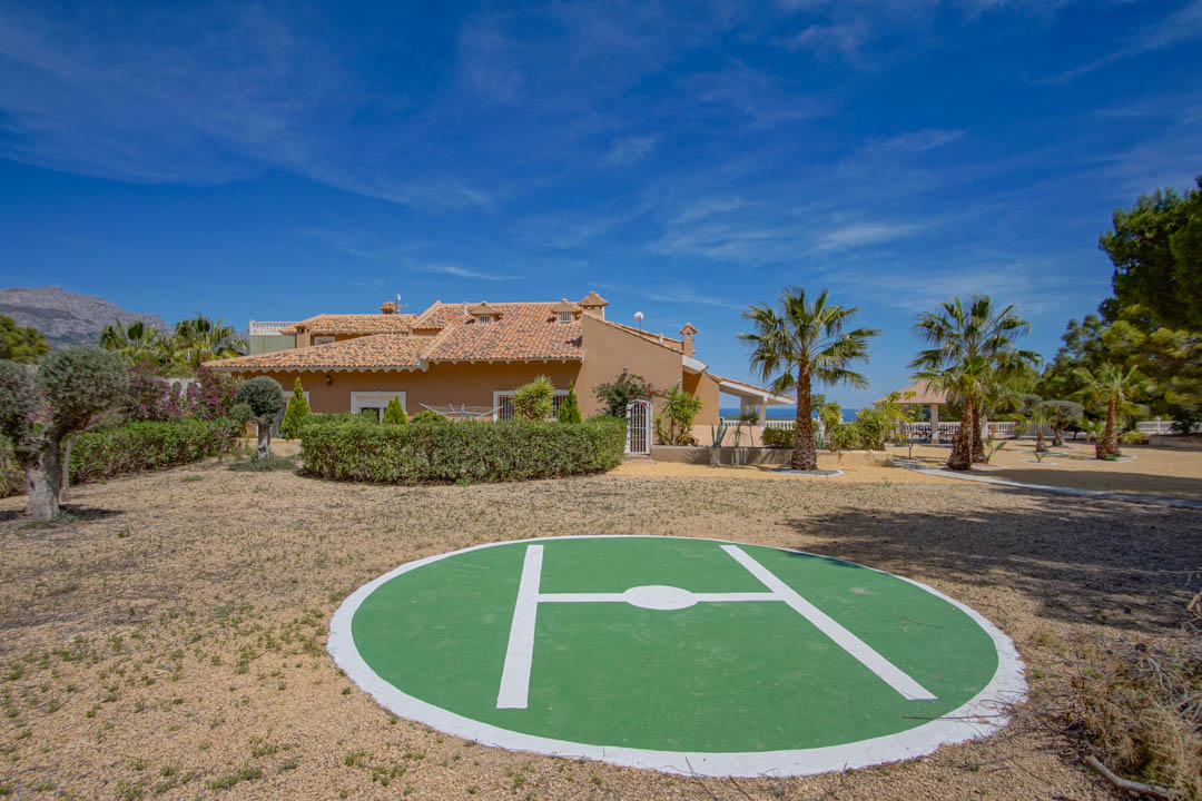 Héliport privé situé à l'arrière de la propriété, entouré d'un jardin méditerranéen avec palmiers et une vue sur la structure traditionnelle de la villa.