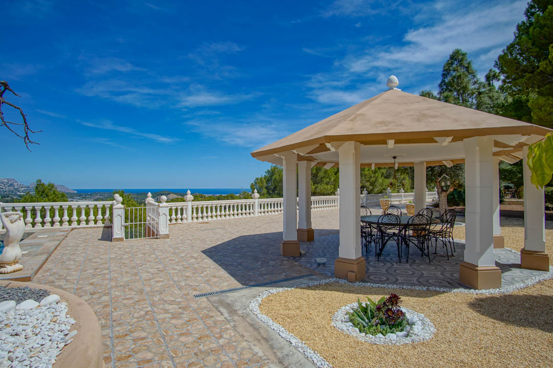 Pavillon extérieur avec colonnes blanches et mobilier en fer forgé, situé sur une terrasse carrelée offrant une vue panoramique sur la mer Méditerranée.