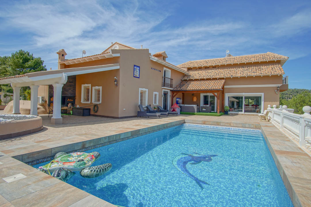 Piscine avec mosaïque décorative, vaste terrasse en pierre et espace barbecue couvert soutenu par des colonnes blanches, reflétant l'architecture méditerranéenne.