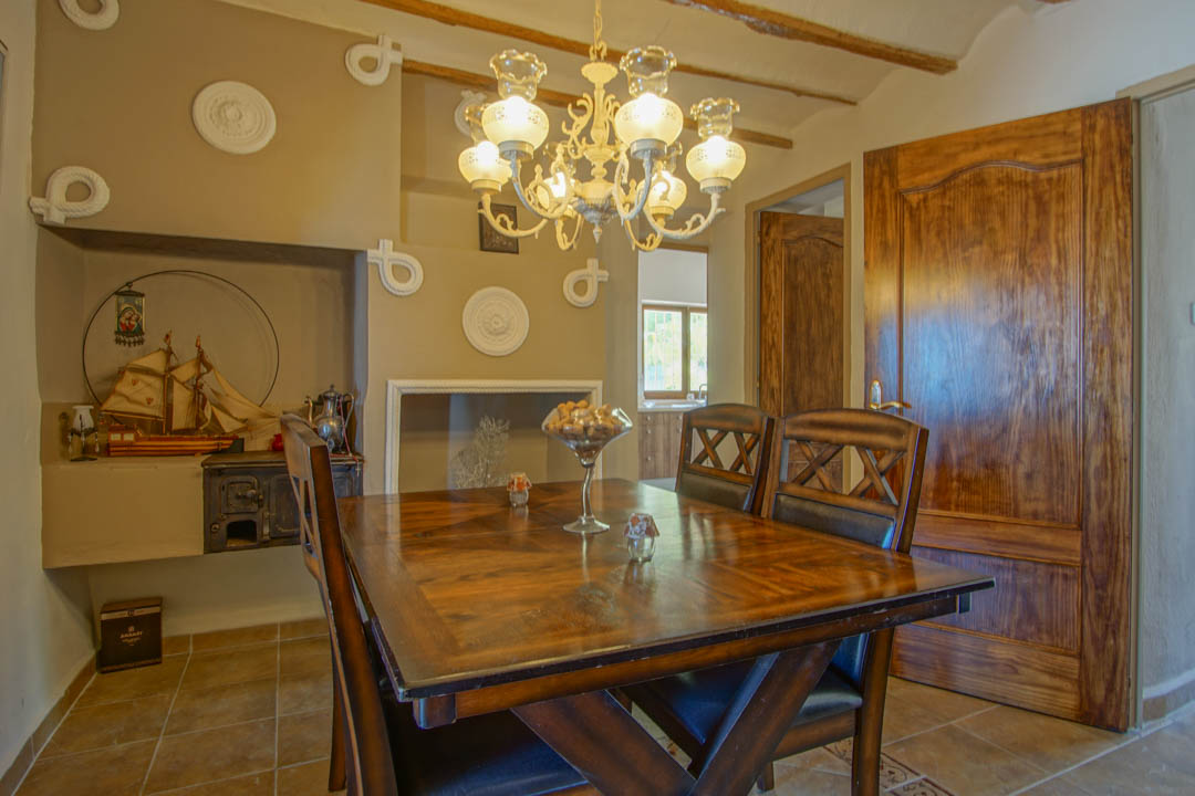 Salle à manger rustique avec table en bois massif, poutres apparentes et lustre classique. Une alcôve traditionnelle abrite un ancien poêle et des décors nautiques.