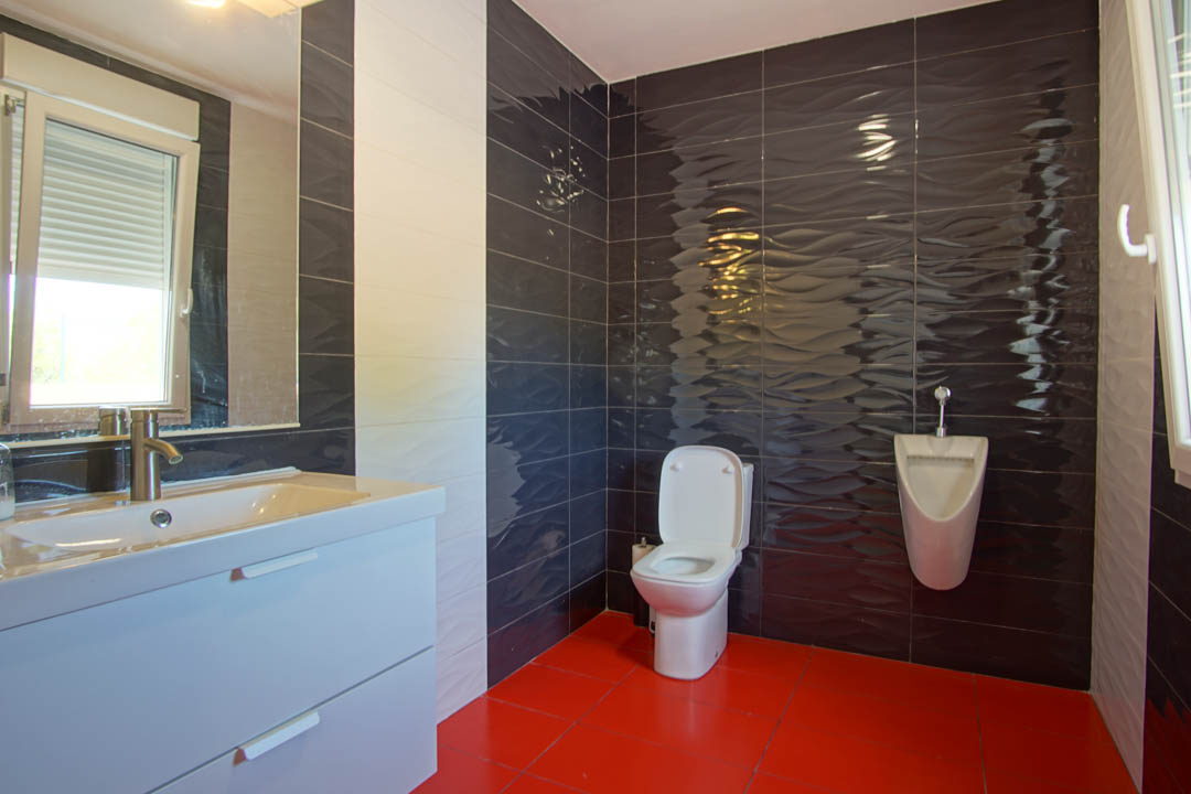 Salle de bain moderne avec carrelage rouge au sol, murs en céramique foncée à texture ondulée, meuble vasque blanc et un urinoir mural.
