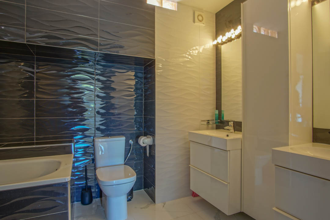 Salle de bain moderne avec carrelage texturé à effet de vagues en blanc et gris foncé, meuble vasque blanc épuré et éclairage intégré au-dessus du miroir.