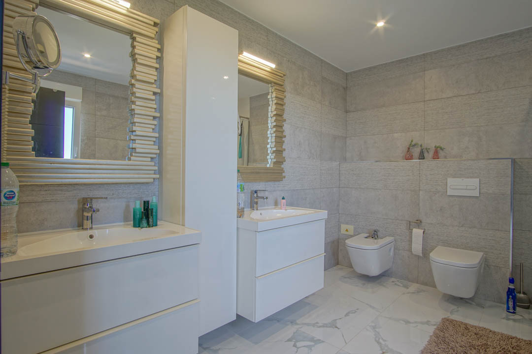 Salle de bain moderne avec double vasque, meubles blancs épurés et miroirs aux cadres texturés. Murs en grès gris et sol en porcelaine effet marbre.
