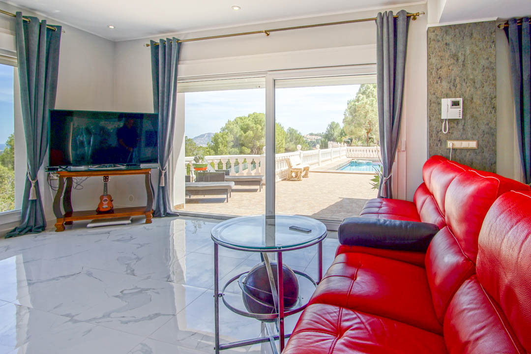 Salon avec sol en marbre blanc, canapé en cuir rouge et grandes baies vitrées s'ouvrant sur une terrasse avec piscine et vue sur la nature.