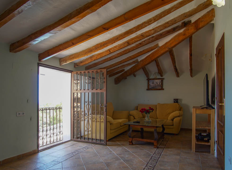 Salon rustique avec poutres en bois apparentes, sol en terre cuite et accès au balcon protégé par une grille décorative en fer forgé.