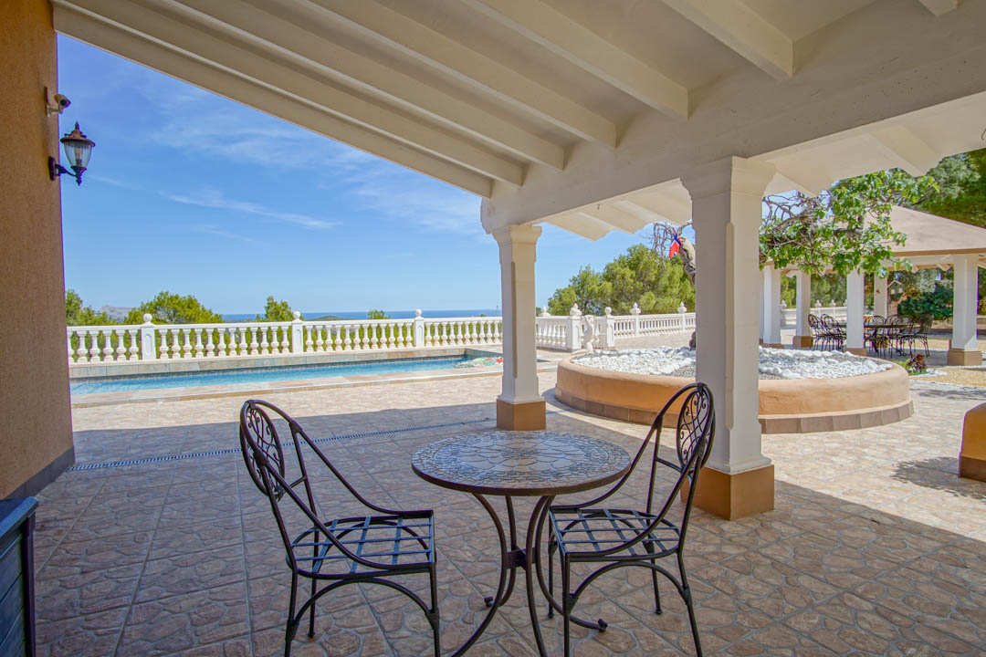 Terrasse couverte avec plafond à poutres blanches, mobilier en fer forgé et vue panoramique sur la piscine et la mer.