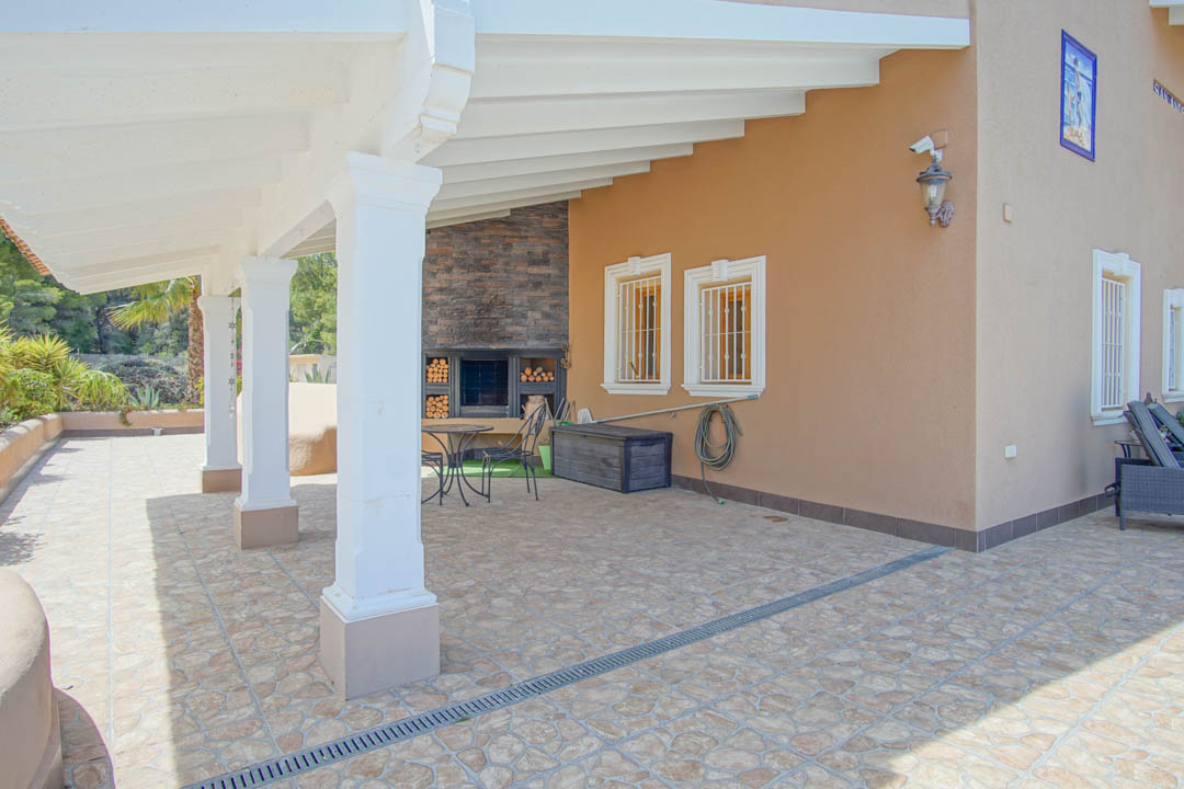 Terrasse couverte avec poutres en bois blanc, colonnes élégantes et cheminée extérieure intégrée en pierre avec espace de rangement pour le bois.