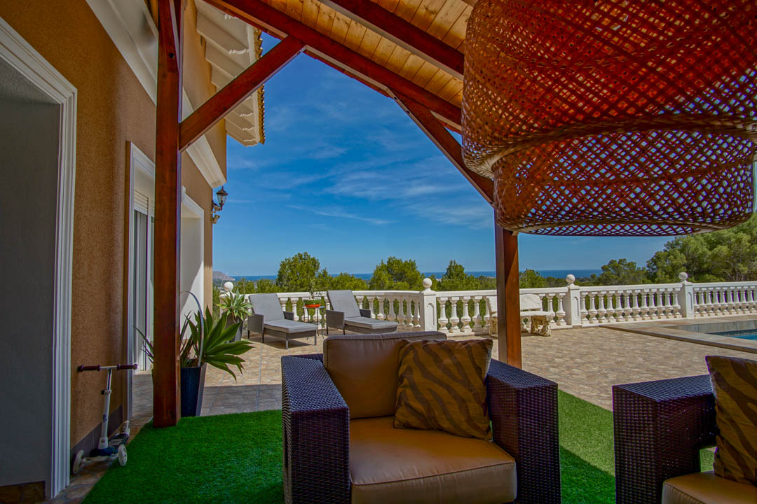 Terrasse couverte avec poutres en bois et salon de jardin, offrant une vue panoramique sur la mer Méditerranée derrière une balustrade blanche.