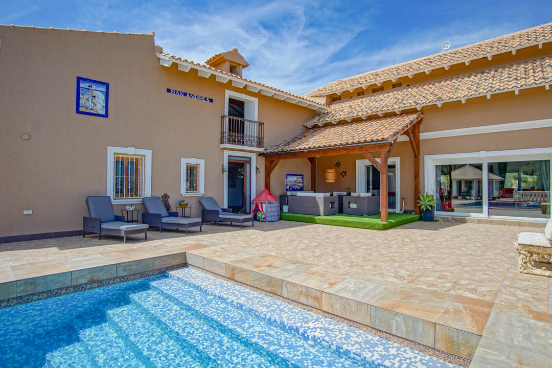 Terrasse principale avec piscine en mosaïque bleue, bains de soleil et porche couvert aux poutres apparentes abritant un salon d'été.