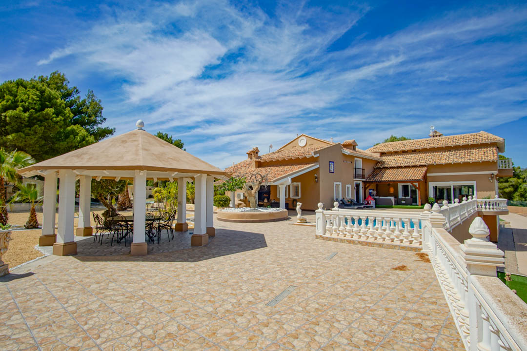 Vaste terrasse extérieure avec dallage en pierre, balustrade classique et pavillon octogonal abritant un coin repas face à la façade de la propriété.