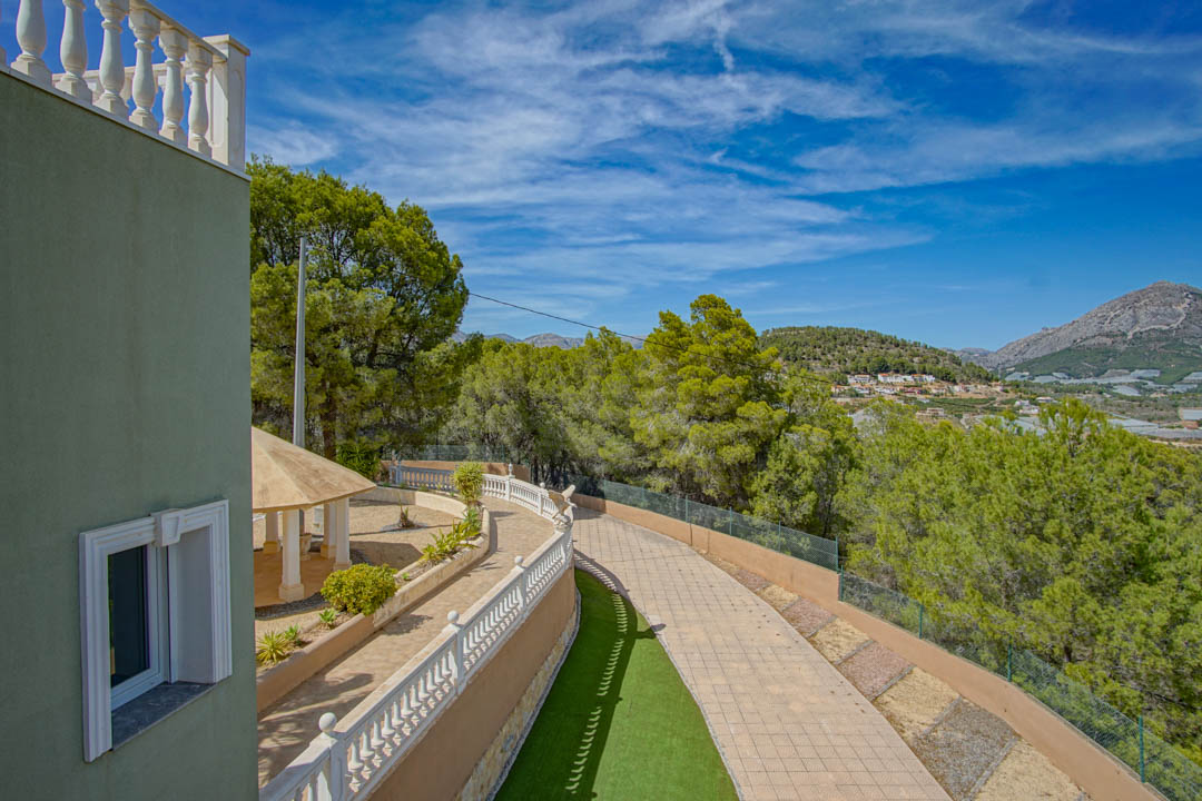Vue sur l'allée pavée et les balustrades blanches. Un pavillon ombragé s'intègre dans le jardin paysager, entouré de pins avec une perspective sur les montagnes.