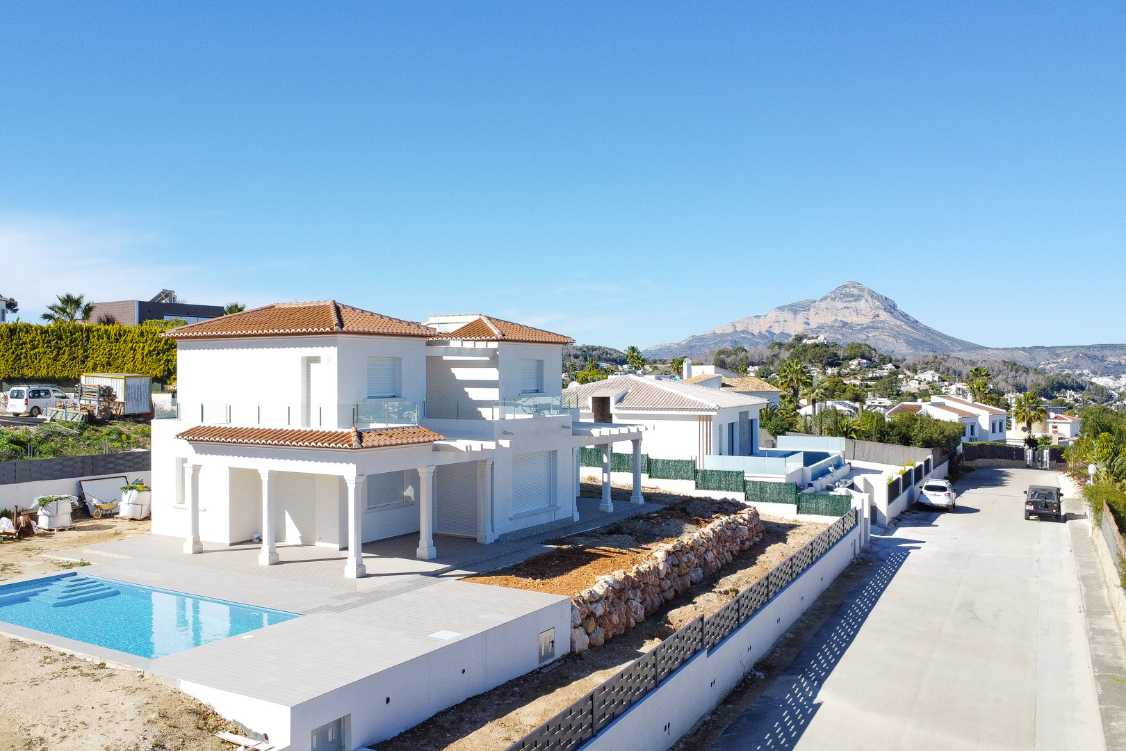 Vue extérieure de la villa blanche avec toit en tuiles, piscine intégrée et murs en pierre naturelle, mettant en valeur le mont Montgó en arrière-plan.